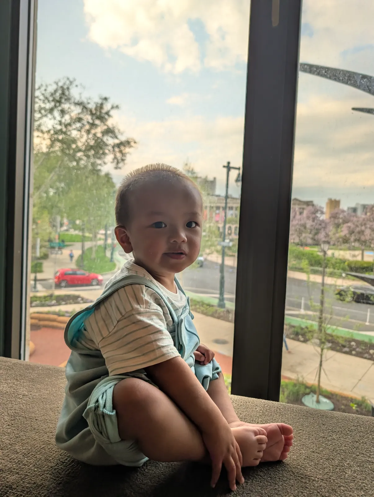 A toddler sitting by a large arched window looking out at the city view