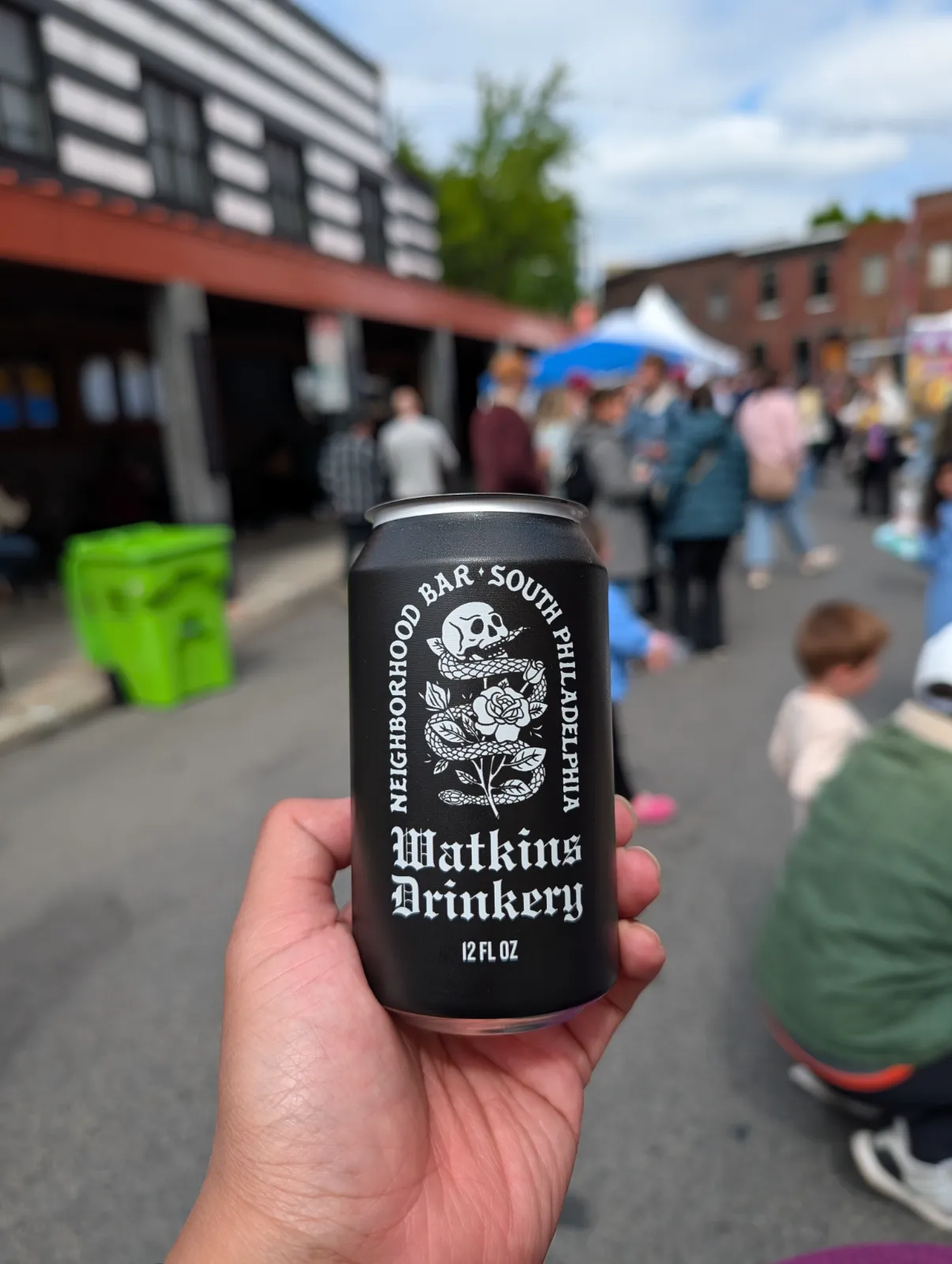 Hand holding a can from Watkins Drinkery with Flavors on the Avenue activity in the background