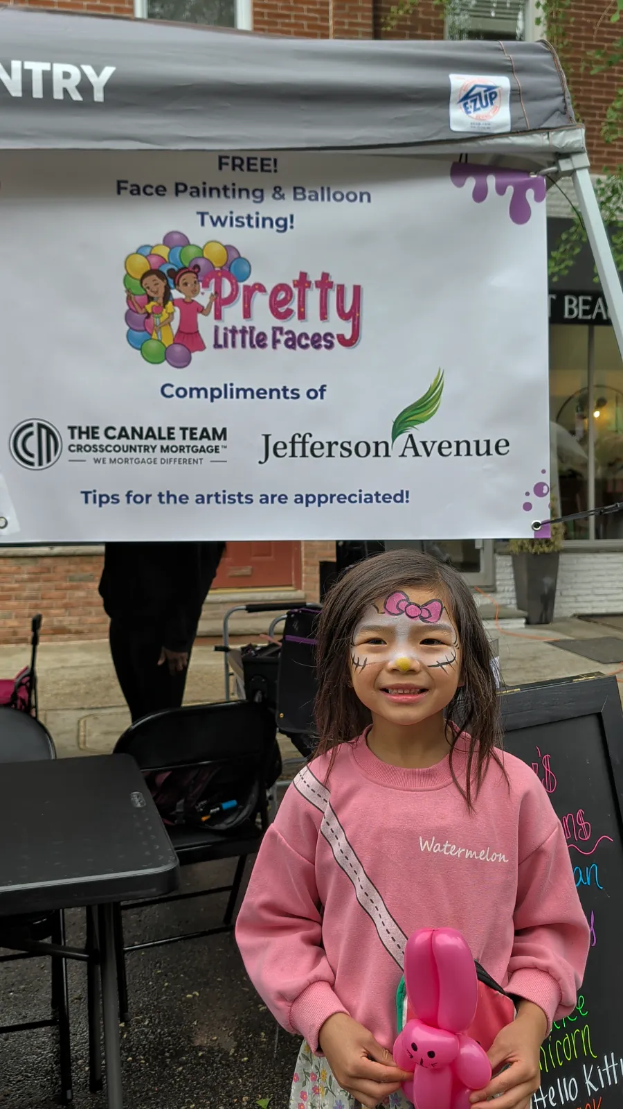 Charlotte holding a pink balloon animal and showing off her face paint at a community event