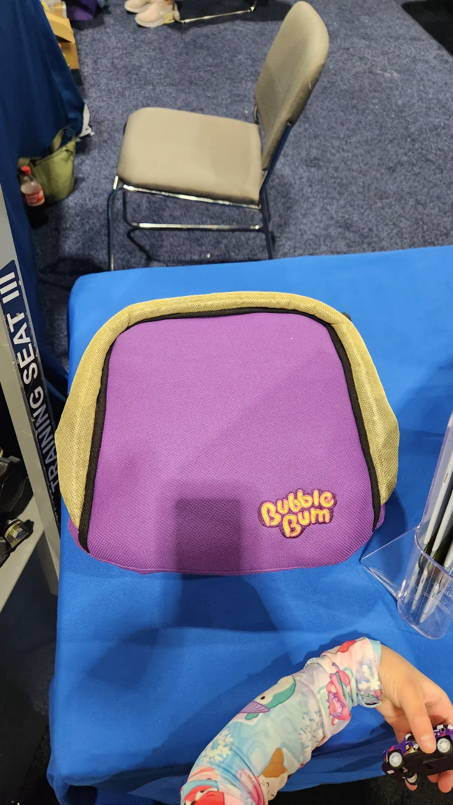 A Bubble Bum portable booster seat bag on display at the Auto Show, with a child reaching for a toy car nearby