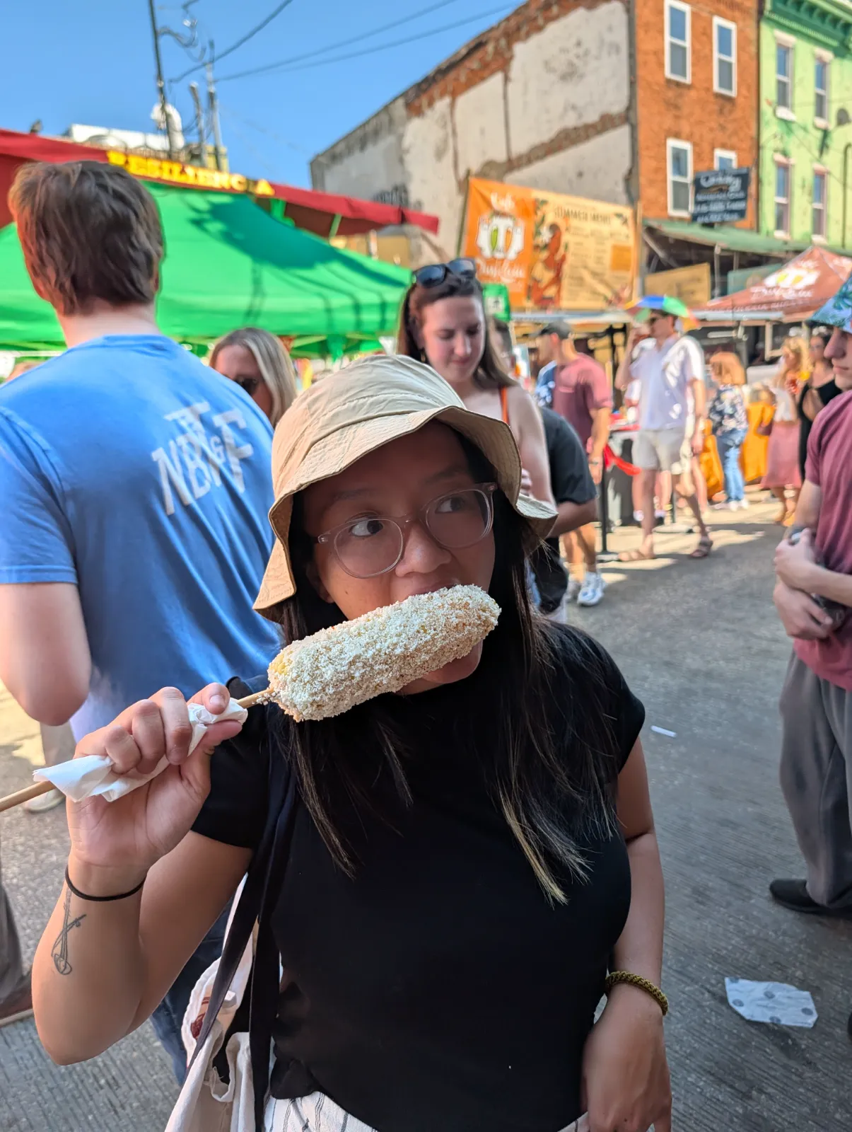 A woman enjoying elote at the Italian Market Festival on 9th Street in South Philadelphia, surrounded by colorful tents and crowds