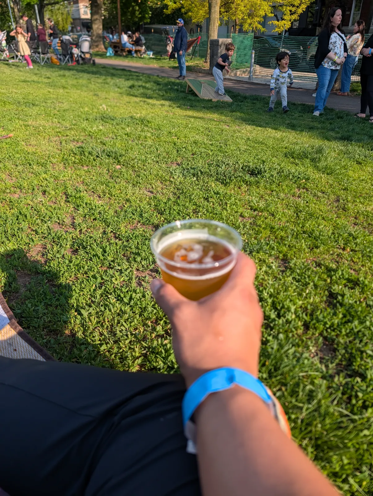 A hand holding a beer at Parks on Tap with families and kids playing in the background
