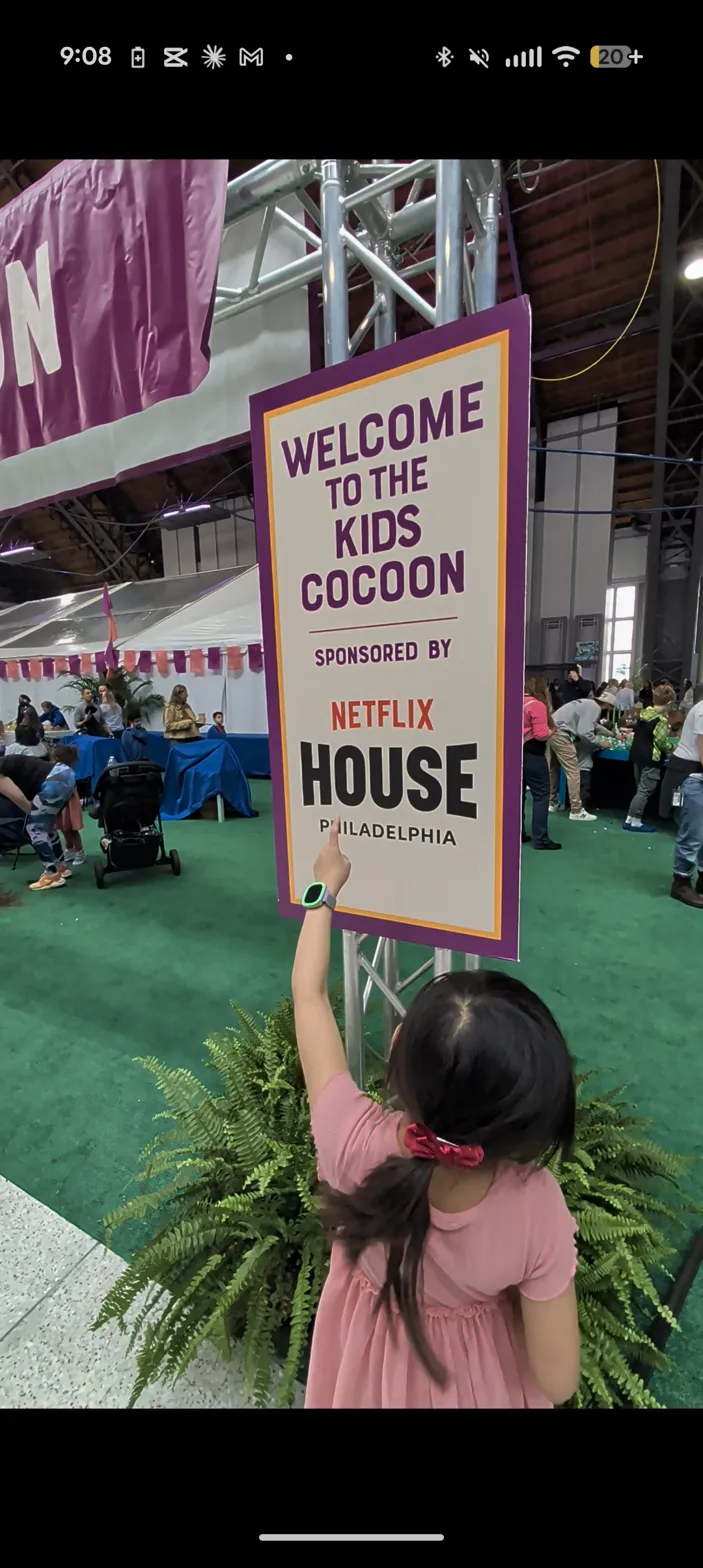 Charlotte reaching up to touch the Welcome to Kids Cocoon sign sponsored by Netflix House Philadelphia
